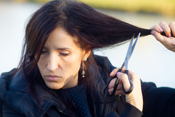 Person cutting their own hair with scissors in a public setting, appearing unaffected by strangers around them.