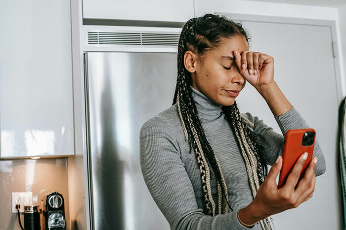 Woman feeling stressed in kitchen while looking at phone, highlighting family and support challenges. Woman feeling stressed in kitchen while looking at phone, highlighting family and support challenges.
