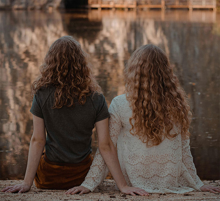 Two women with curly hair sit by a lake, facing away, symbolizing distance and family expectations. Two women with curly hair sit by a lake, facing away, symbolizing distance and family expectations.