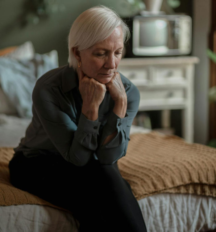 Elderly woman sitting on bed looking pensive, related to a hyphenated last name discussion in a family setting. Elderly woman sitting on bed looking pensive, related to a hyphenated last name discussion in a family setting.