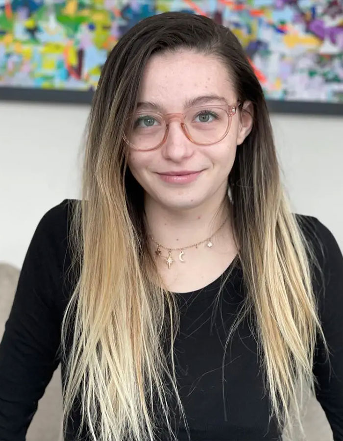 Young woman with long blond hair and glasses, wearing a black top, seated indoors with a colorful painting in background. Young woman with long blond hair and glasses, wearing a black top, seated indoors with a colorful painting in background.