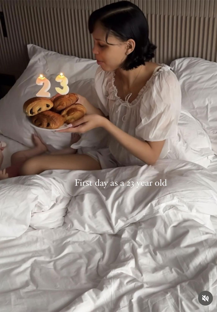 Nara Smith in bed holding pastries with lit number 23 candles, calm and composed in a white dress.