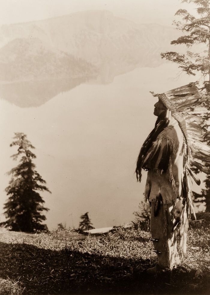 Native American man in traditional headdress standing by a lakeside overlooking mountains in a haunting historic photo.