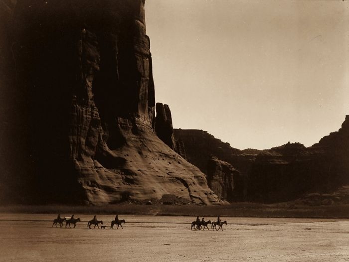 Native Americans on horseback crossing a vast desert landscape with towering rock formations in the background.