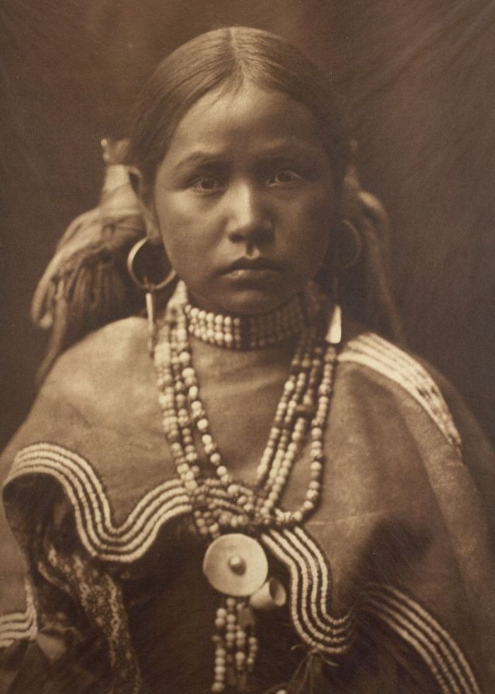 Young Native American girl wearing traditional beaded jewelry and clothing in a haunting historical photo from a century ago.