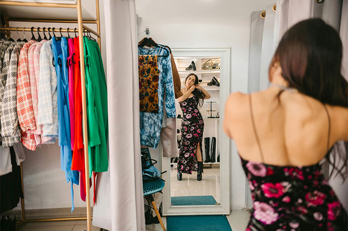 Woman trying on a dress in a store, looking at her reflection, amidst colorful clothes, reflects no-buy challenge mindset. Woman trying on a dress in a store, looking at her reflection, amidst colorful clothes, reflects no-buy challenge mindset.