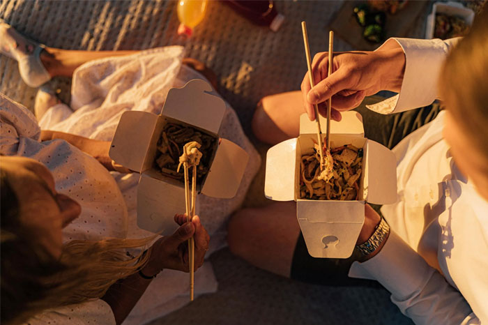 Two people enjoying takeout with chopsticks, possibly embracing a "No-Buy" lifestyle to resist consumerism. Two people enjoying takeout with chopsticks, possibly embracing a "No-Buy" lifestyle to resist consumerism.
