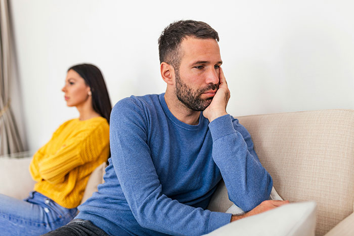 Man in a blue sweater sitting on a couch, looking pensive; woman in a yellow sweater sits back-to-back. Man in a blue sweater sitting on a couch, looking pensive; woman in a yellow sweater sits back-to-back.