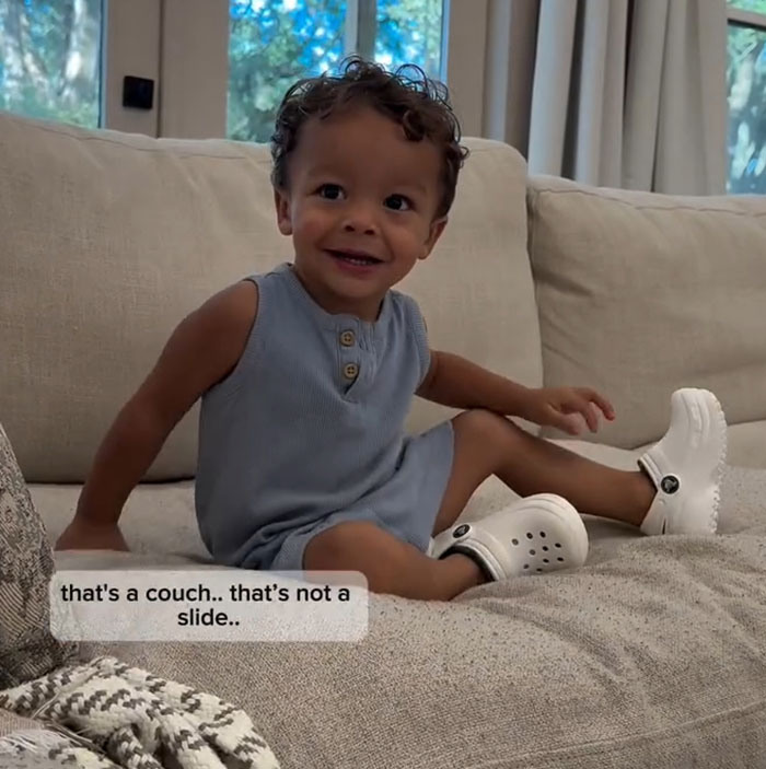 Toddler from 'Okay Baby' smiling on a couch, wearing a blue outfit and white shoes. Toddler from 'Okay Baby' smiling on a couch, wearing a blue outfit and white shoes.