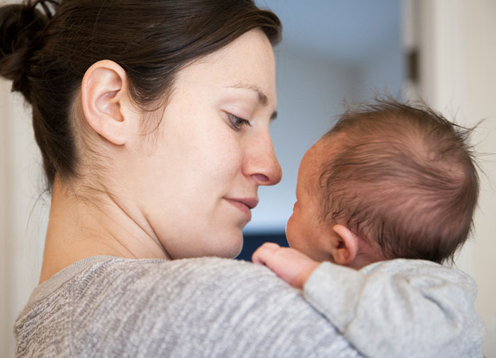 New mom holding her newborn, facing away from the camera in a softly lit room. New mom holding her newborn, facing away from the camera in a softly lit room.