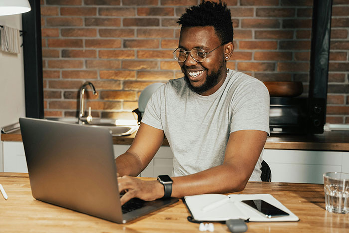 Man smiling at laptop in kitchen, planning an expensive trip to visit long-distance girlfriend. Man smiling at laptop in kitchen, planning an expensive trip to visit long-distance girlfriend.