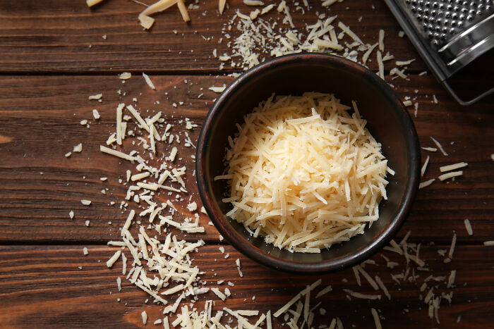 Bowl of grated cheese with scattered shreds on wooden surface, illustrating usual things altered by the pandemic.