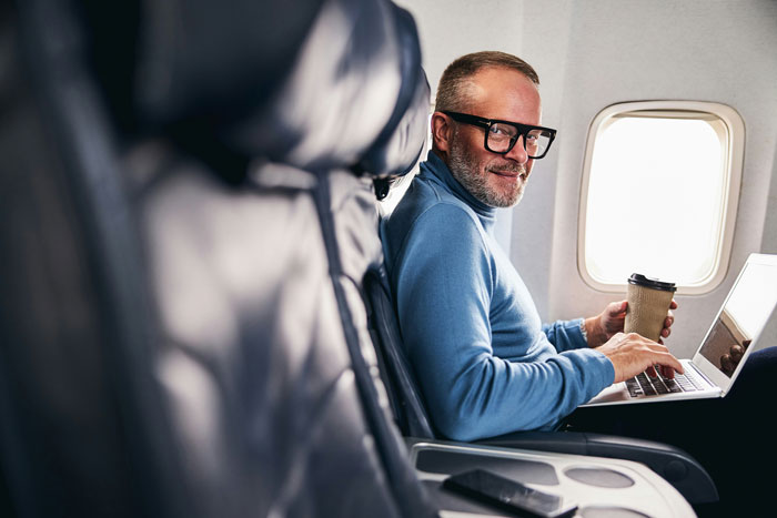 Man in airplane seat with coffee and laptop, smiling, embodying a relaxed passenger experience. Man in airplane seat with coffee and laptop, smiling, embodying a relaxed passenger experience.