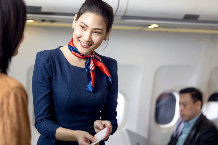 Flight attendant smiling while handling ticket, passenger reading nearby; highlighting travel experience. Flight attendant smiling while handling ticket, passenger reading nearby; highlighting travel experience.