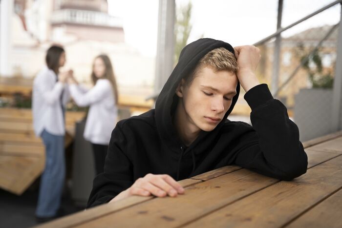 A young person in a hoodie looks down, with two people talking in the background, illustrating bullying issues.