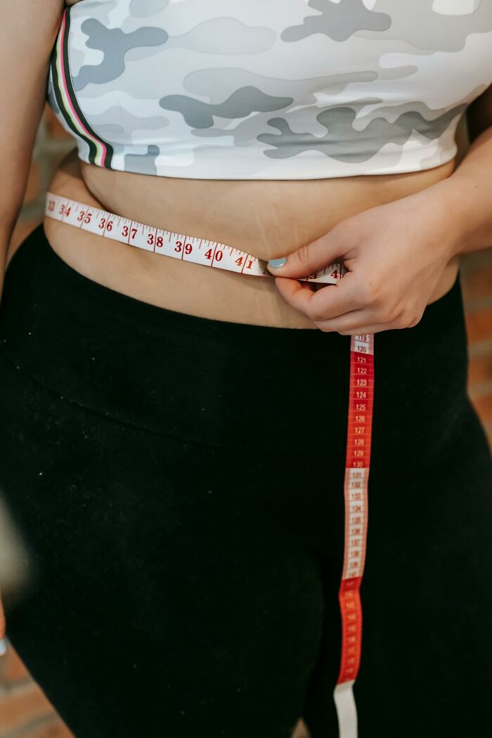 Woman measuring waist with tape measure, wearing a camo top and black pants, illustrating body image concerns.