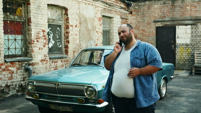 A man stands near a vintage car in an urban alley, appearing thoughtful.