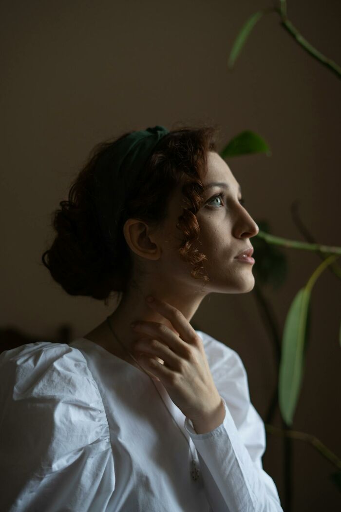 Woman in white shirt looking pensive, with hand at her neck, surrounded by plants; exploring women's embarrassment.