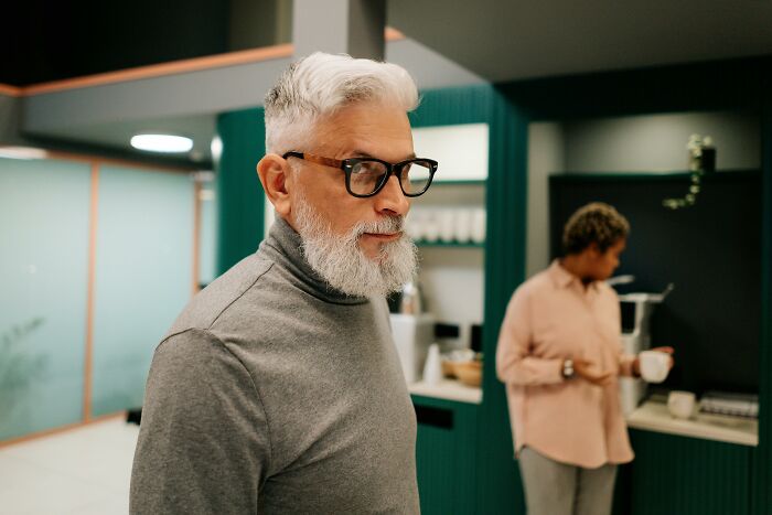 Man with gray hair and glasses in a modern kitchen, with a person in the background.