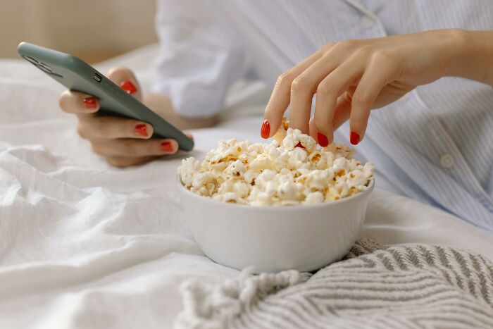 Person with red nails holds a phone and eats popcorn from a bowl, searching for a movie. Person with red nails holds a phone and eats popcorn from a bowl, searching for a movie.
