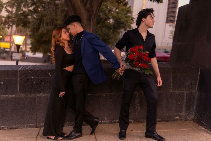 Three young adults showing unexplainable pain, two embracing and one holding roses walking away in a public park.