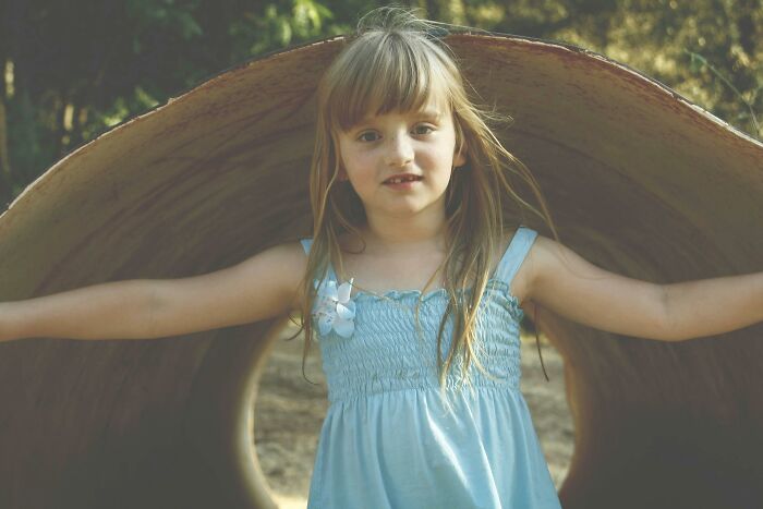 Young girl in a blue dress standing inside a large wooden tunnel, capturing a moment outdoors and innocence.