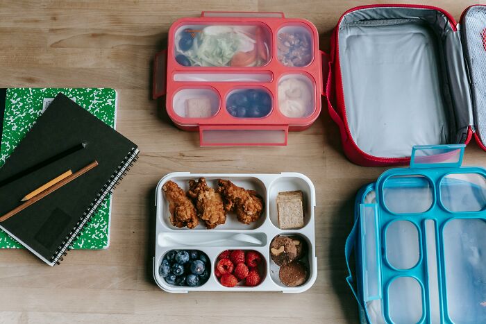 Two lunchboxes with fried chicken, berries, and notebooks on a wooden table; concept of sharing food experiences.