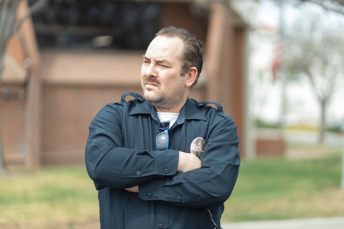 Man in a black shirt with crossed arms outdoors, appearing skeptical in a park setting.