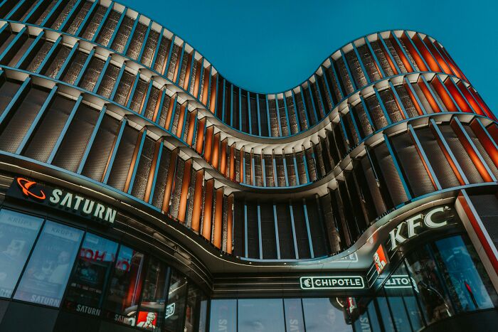 Curved building facade with Saturn, Chipotle, and KFC logos, glowing under evening sky.