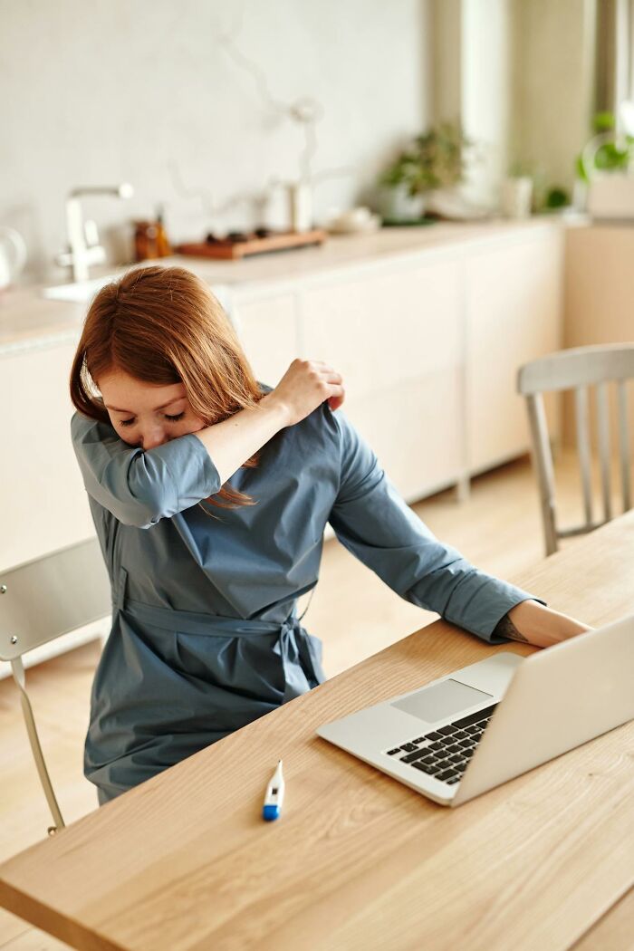 Woman sneezing into elbow at table, with laptop and thermometer, capturing a moment that may puzzle men.