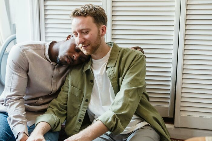 Two men sitting close together indoors, one resting his head on the other's shoulder, conveying comfort and support.