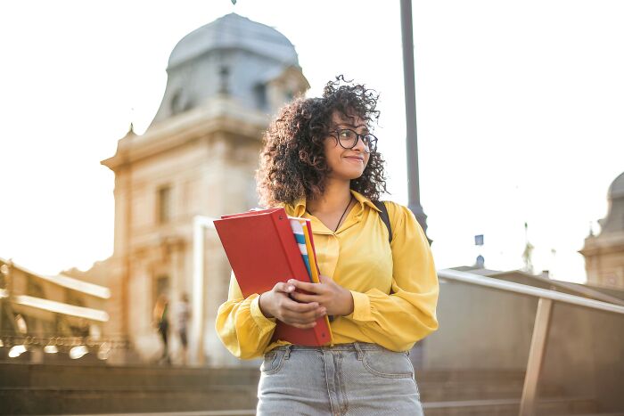 Person with books on campus, wearing a yellow shirt and glasses, smiling in front of a historic building.