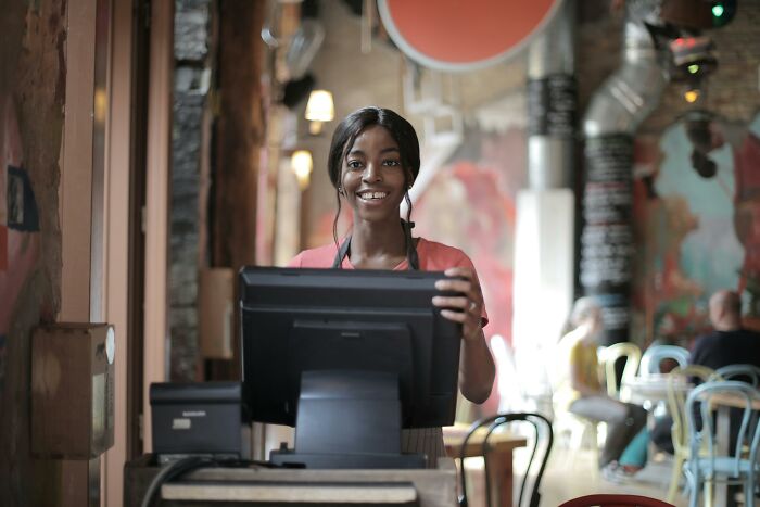 Smiling woman at a cafe register, standing ready to assist customers amidst colorful decor, possibly avoiding any adult tantrums.