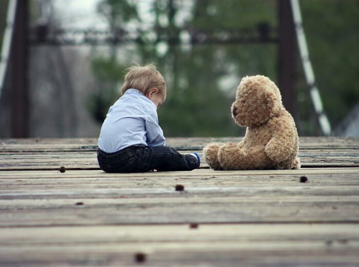Young child sitting on a wooden bridge facing a teddy bear, symbolizing unexplainable pain endured alone.