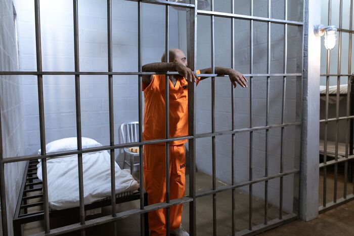 Man in orange prison jumpsuit leaning on jail bars, appearing thoughtful.