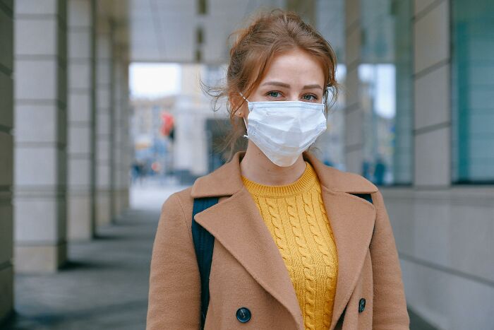 Woman in a tan coat and yellow sweater wearing a mask, standing confidently in an urban environment.