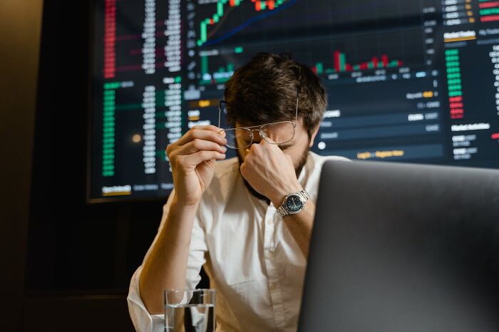 Man in white shirt, sitting at a computer, looks stressed with hand on face, stock market graphs in background, April 1st.