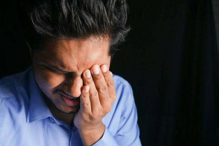 Man in blue shirt enduring unexplainable pain, holding face with eyes closed against a dark background