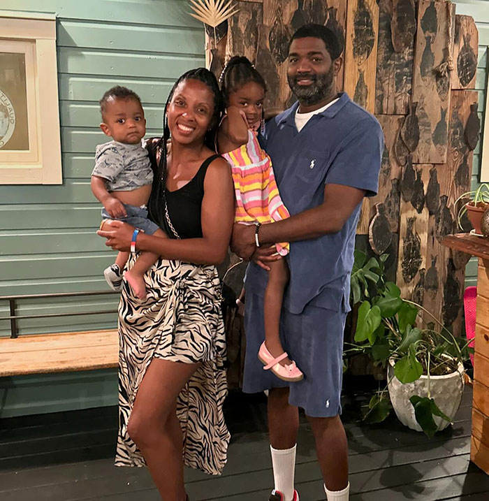 A family of four smiling inside a wooden-themed room, with two adults holding kids, during a casual photo shoot. A family of four smiling inside a wooden-themed room, with two adults holding kids, during a casual photo shoot.