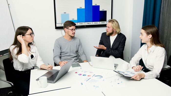A team of four in a meeting, discussing productivity tips with charts on a monitor in the background.