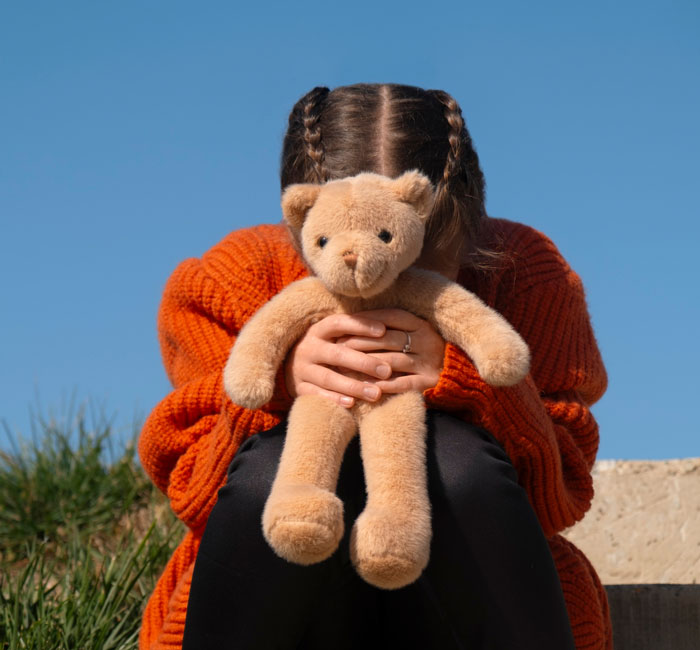 Girl with leukemia holding teddy bear tightly, sitting outdoors in orange sweater on a sunny day.