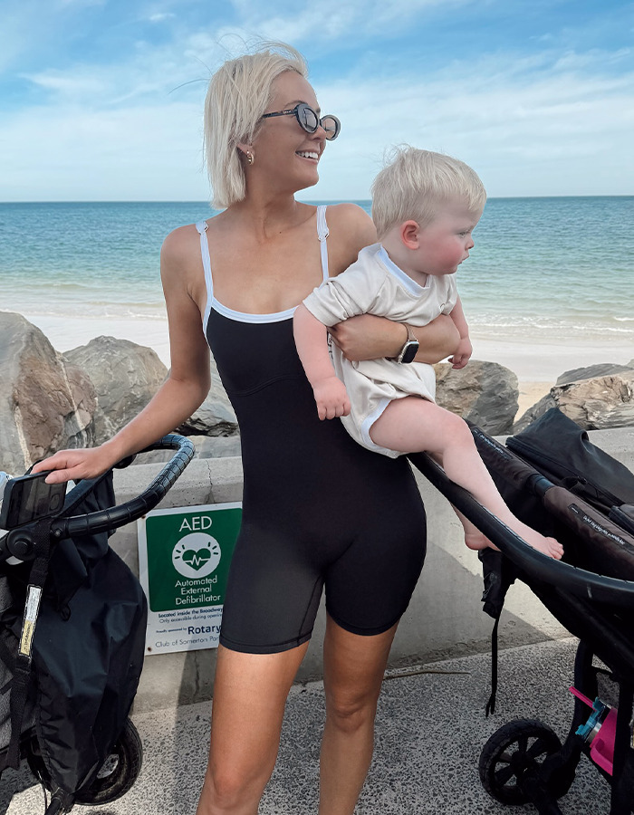 Woman holding a toddler near the beach, surrounded by strollers, with a cheerful expression. Woman holding a toddler near the beach, surrounded by strollers, with a cheerful expression.