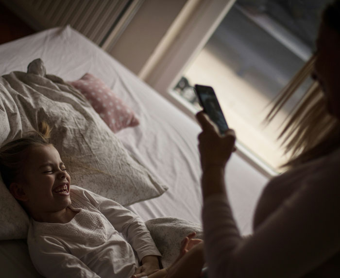A parent photographs their daughter in bed, smiling during an intimate moment, highlighting personal sharing online. A parent photographs their daughter in bed, smiling during an intimate moment, highlighting personal sharing online.