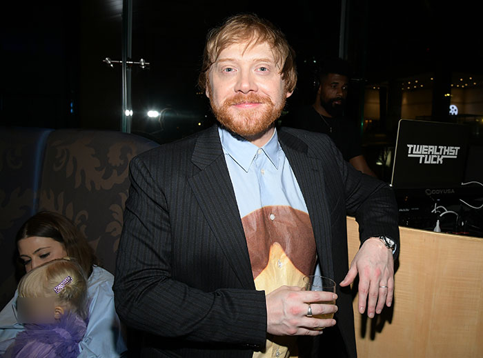 Rupert Grint, smiling at an event, with family in the background. Rupert Grint, smiling at an event, with family in the background.
