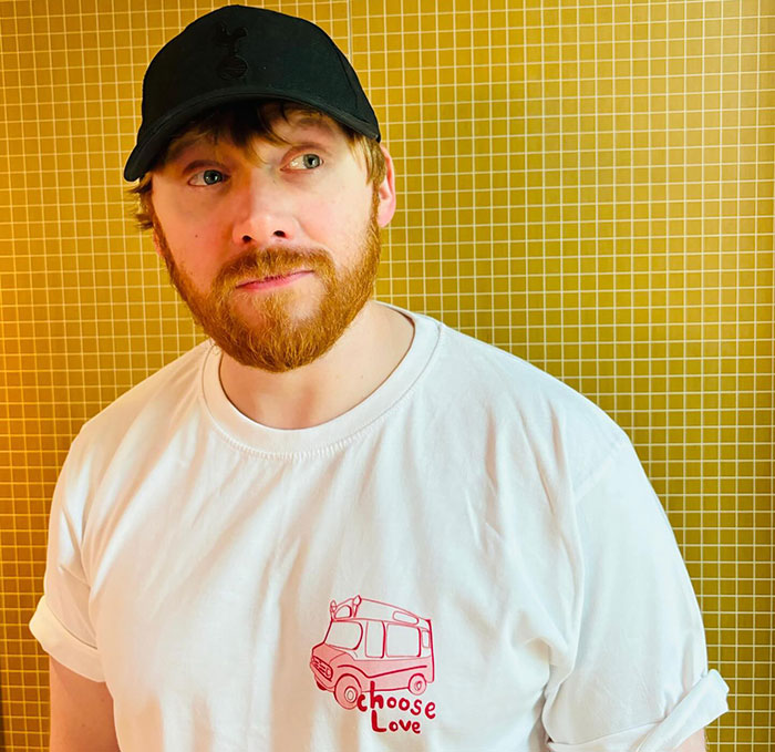 Actor in a cap and "Choose Love" shirt against a yellow tiled background. Actor in a cap and "Choose Love" shirt against a yellow tiled background.