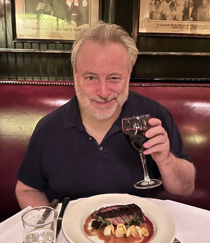 Man in black shirt holding a wine glass at a restaurant, with a dish in front of him, referencing punk rock legend Patti Smith. Man in black shirt holding a wine glass at a restaurant, with a dish in front of him, referencing punk rock legend Patti Smith.