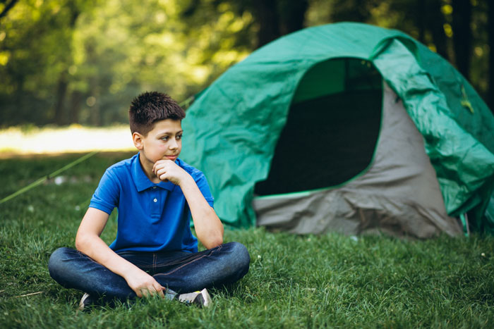 Boy in a blue shirt sitting on grass beside a green tent, outdoors in a park setting. Boy in a blue shirt sitting on grass beside a green tent, outdoors in a park setting.