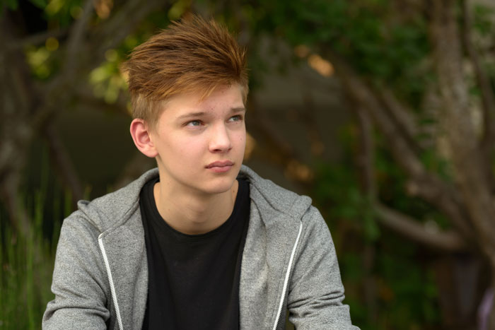 Teen boy in gray hoodie sitting outdoors, illustrating dad catching teen son creeping around neighbor’s yard at night. Teen boy in gray hoodie sitting outdoors, illustrating dad catching teen son creeping around neighbor’s yard at night.