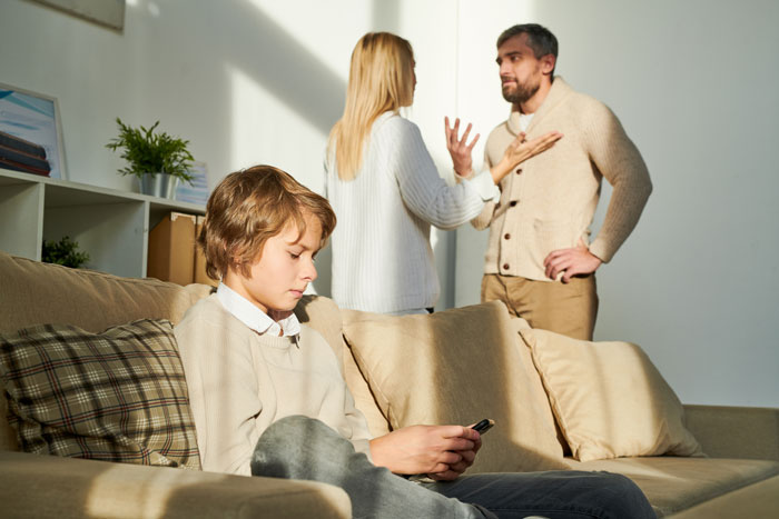 Son sitting on a couch distracted by phone while father and mother argue in the background, highlighting son father grooming drama. Son sitting on a couch distracted by phone while father and mother argue in the background, highlighting son father grooming drama.