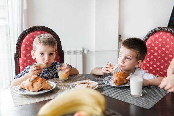Two boys sitting at a table, eating breakfast with juice and milk, related to allergies and family concerns. Two boys sitting at a table, eating breakfast with juice and milk, related to allergies and family concerns.